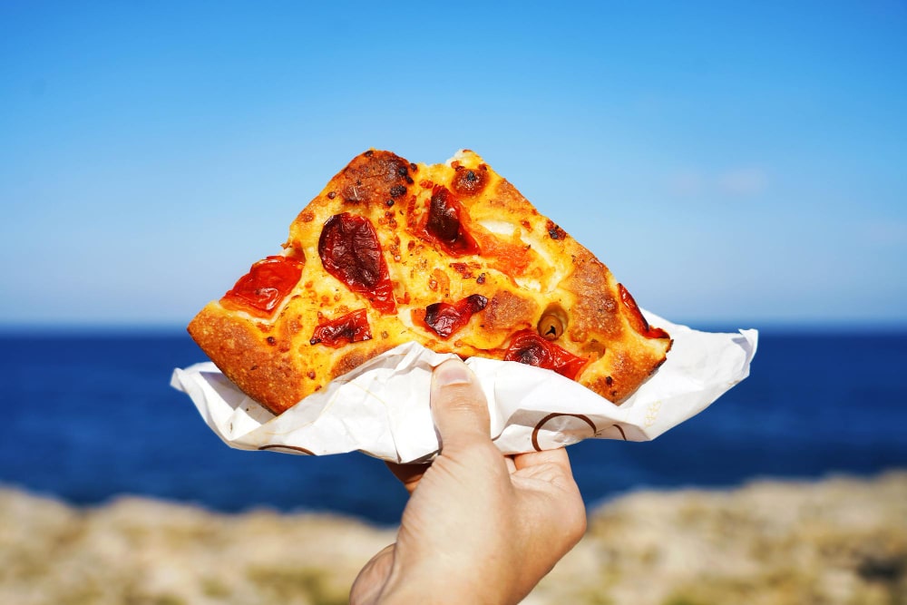 focaccia-barese-slice-with-mediterranean-sea-background-focaccia-pugliese-against-blue-sea-sky-baristyle-focaccia-bread-min